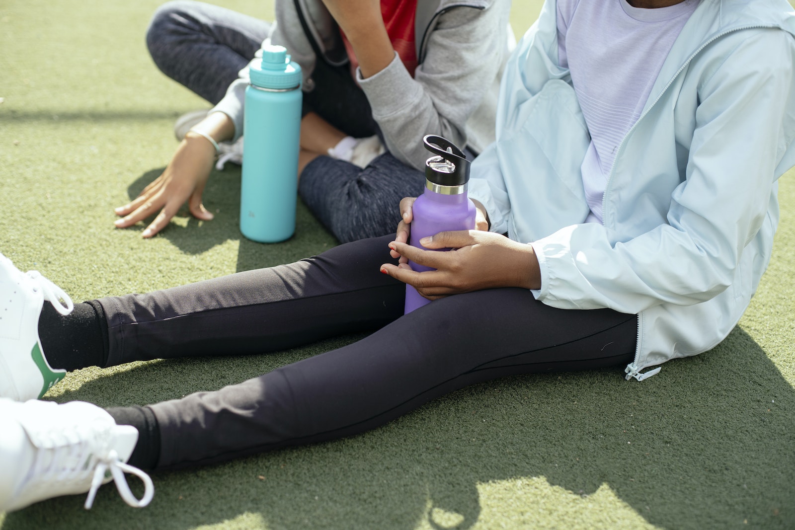 Crop schoolgirls with plastic water bottles sitting on artificial grass field while having break in training