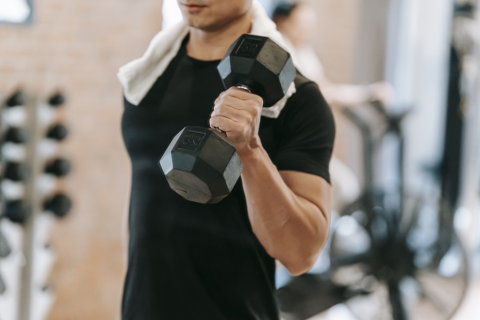 Unrecognizable strong male with towel wearing black activewear lifting heavy dumbbell during intense training in fitness studio with special equipment on blurred background