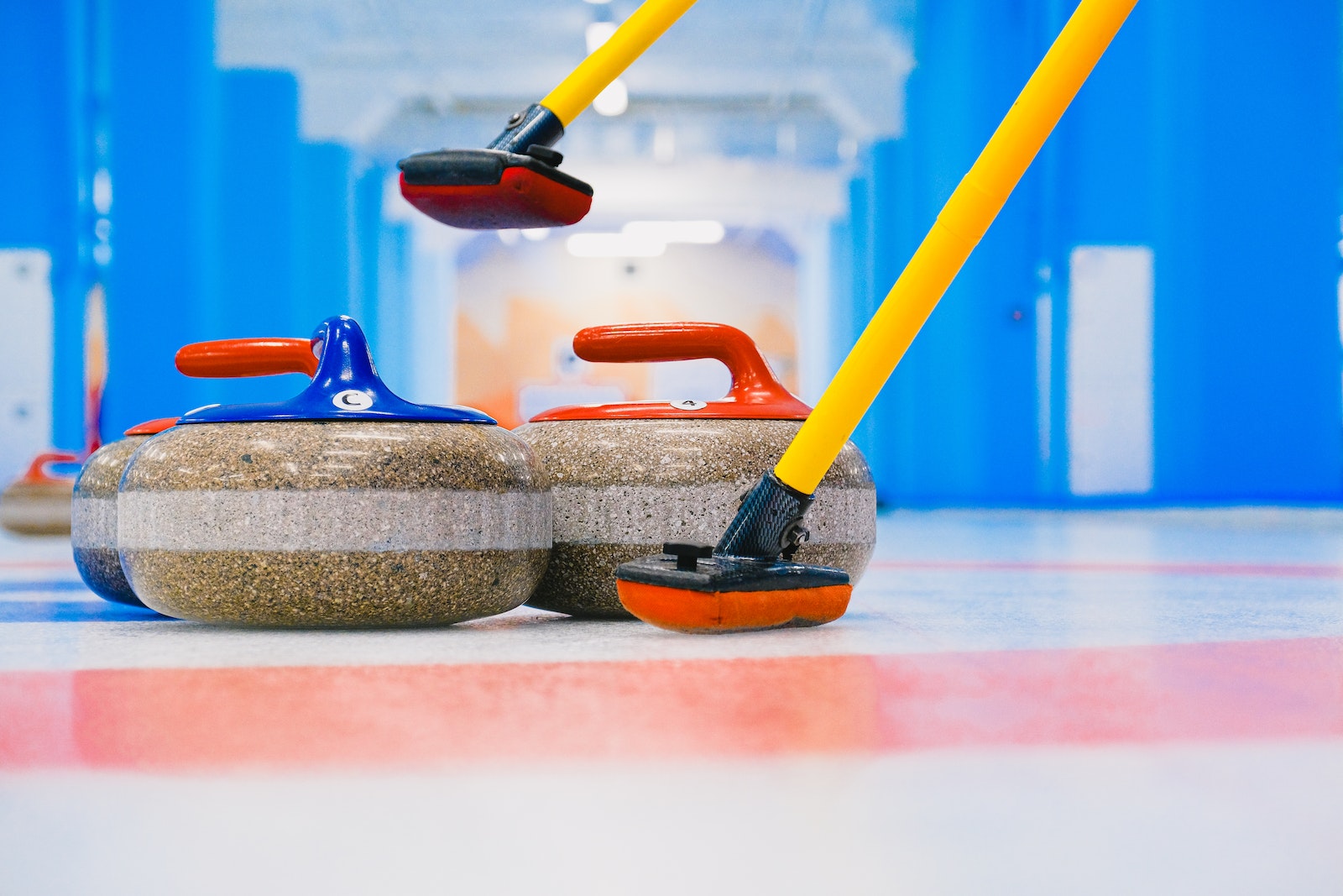 Granite curling stones with handles and long brooms placed on circular target area on ice sheet in arena with blue columns
